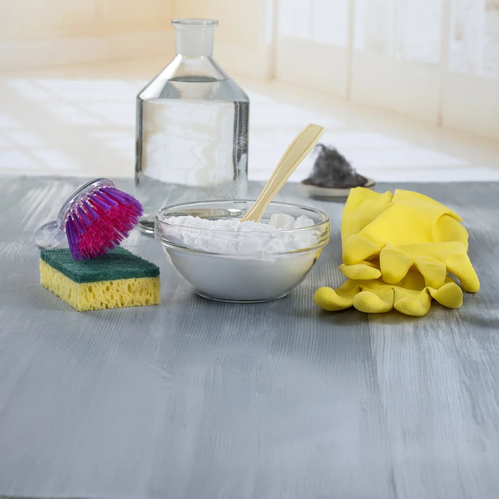 Reinigungsutensilien auf einem Tisch: Eine Schüssel mit Natron und einem Löffel, eine Flasche Essig, ein gelber Reinigungshandschuh, ein Schwamm mit einer Bürste mit pinken Borsten sowie ein kleiner Haufen Stahlwolle im Hintergrund.
