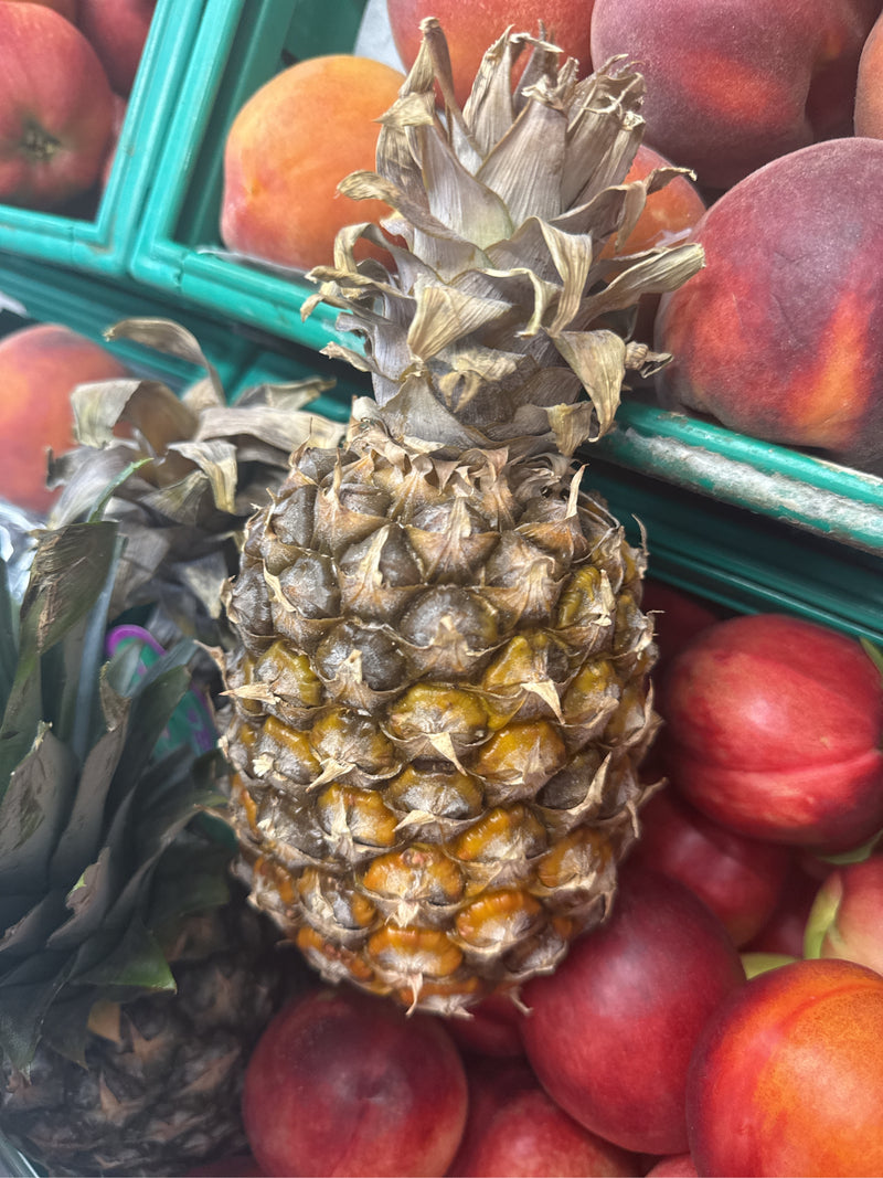 Zu sehen ist eine reife Ananas mit schuppiger Haut und grünem Schopf, die zwischen roten Früchten in einer Auslage liegt.