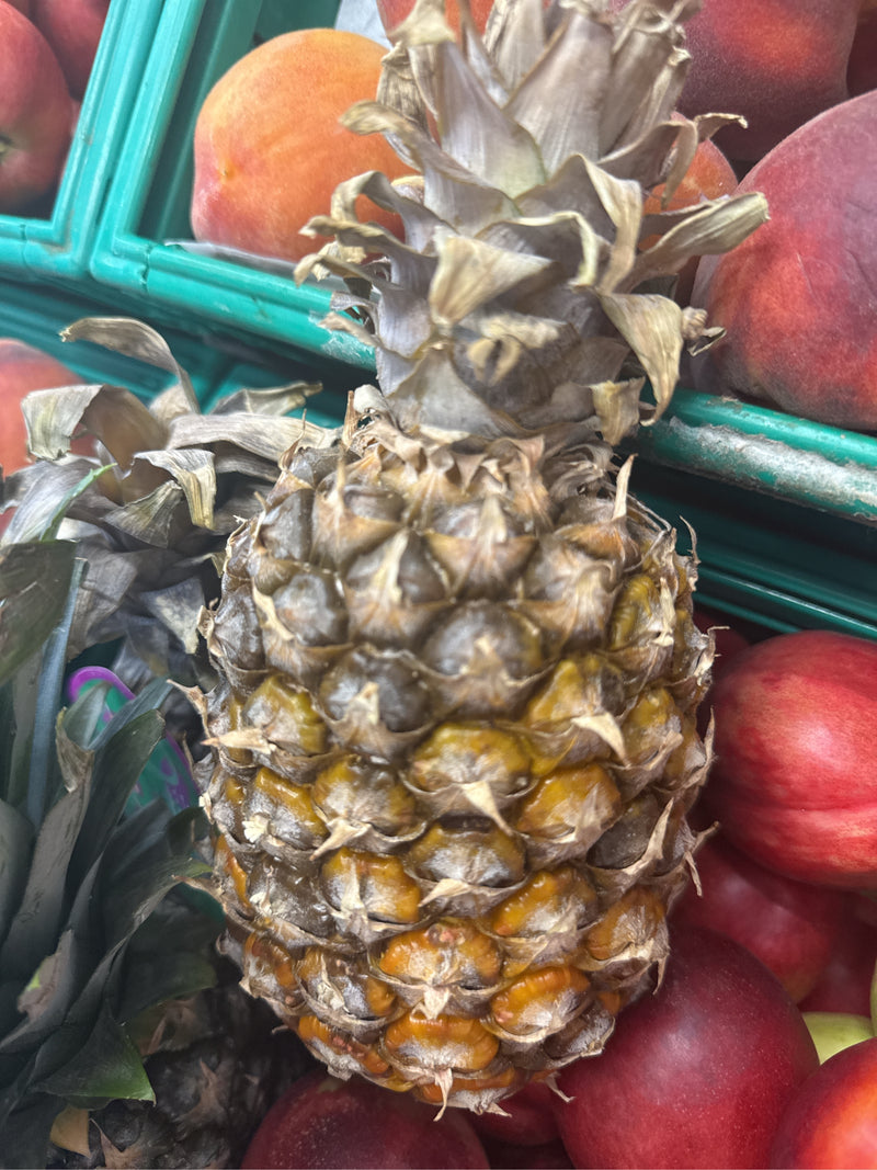 Auf dem Bild ist eine reife Ananas mit ihrem charakteristischen Blattschopf zu sehen, platziert zwischen anderem Obst in einer Auslage.