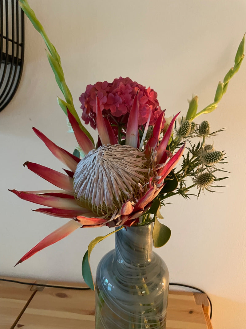Zu sehen ist eine grosse Blüte der Königsprotea (Protea cynaroides), die Teil eines bunten Blumenstrausses in einer Glasvase ist. Die markanten, spitz zulauf...