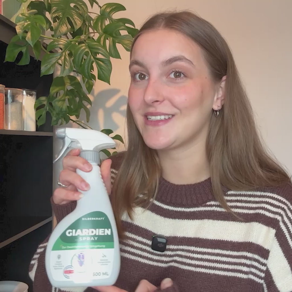 Ein Werbevideo der Marke Silberkraft für ein Giardien-Spray zur Desinfektion der Umgebung von Haustieren. Eine junge Frau erklärt die Anwendung: Sie schüttelt eine weiße Sprühflasche und besprüht einen grauen Teppich. Das Video zeigt zudem Ausschnitte von einem Hund, einer Katze am Futternapf sowie Infografiken zu Einsatzbereichen wie Tierbetten und Katzenklos. Ein Textoverlay hebt Vorteile wie „Wirksam gegen Giardien“, „OHNE Permethrin“ und „Qualität aus Deutschland“ hervor.